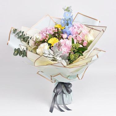 Birthday Happiness, Blooms of Joyful Celebration Bouquet, Light Pink Hydrangea, White Ohara Garden Rose, Yellow Gerbera, Light Blue Delphinium, Green Skimmia Filler, Cinerea Eucalyptus Filler, Light Pink Wax Filler, Casino Pink Hypericum Coco Filler, Arranged in a Bouquet
